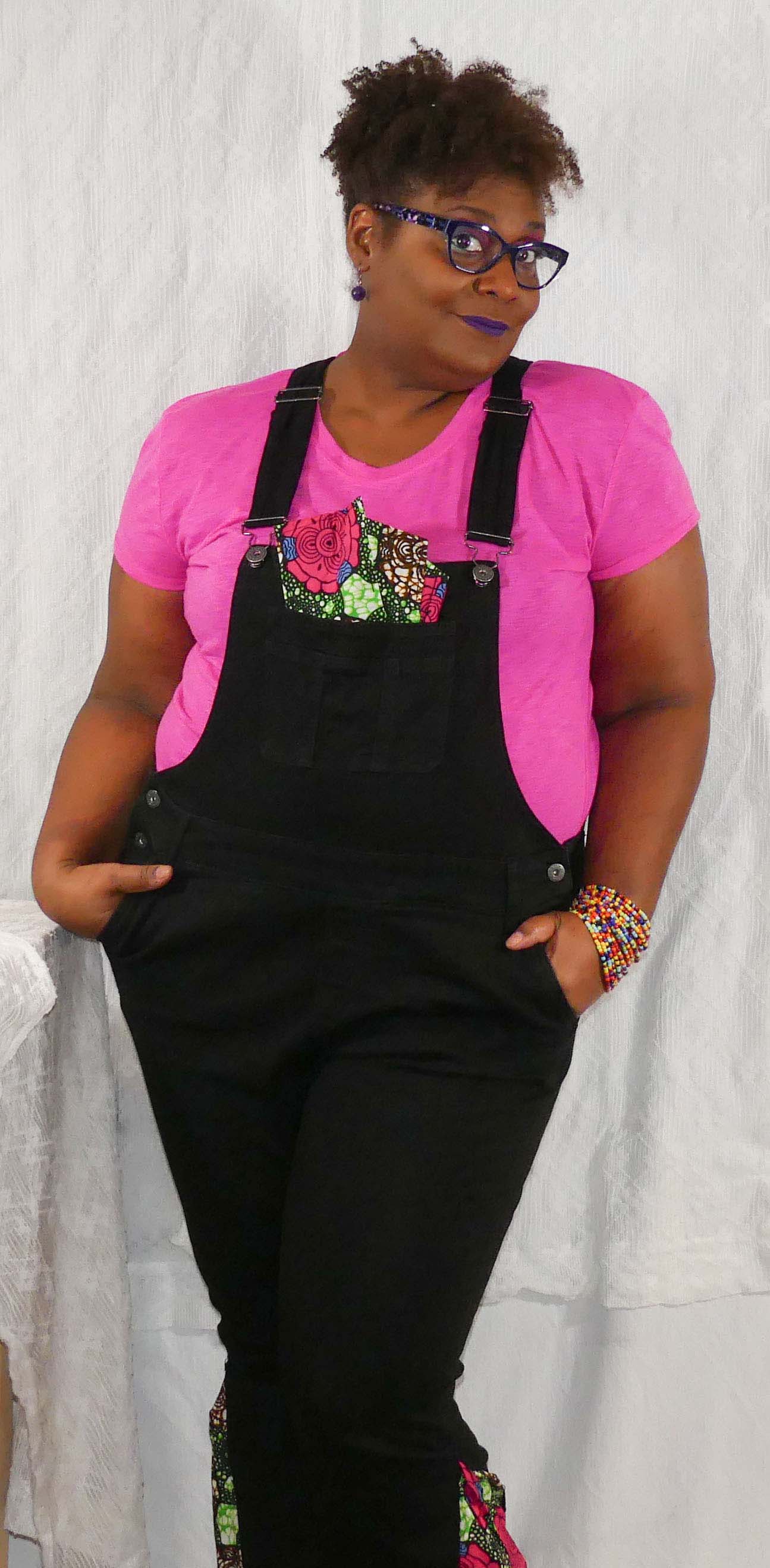 Sonya wearing a pink t-shirt and black overalls with a colorful pocket design, standing against a light-colored backdrop.